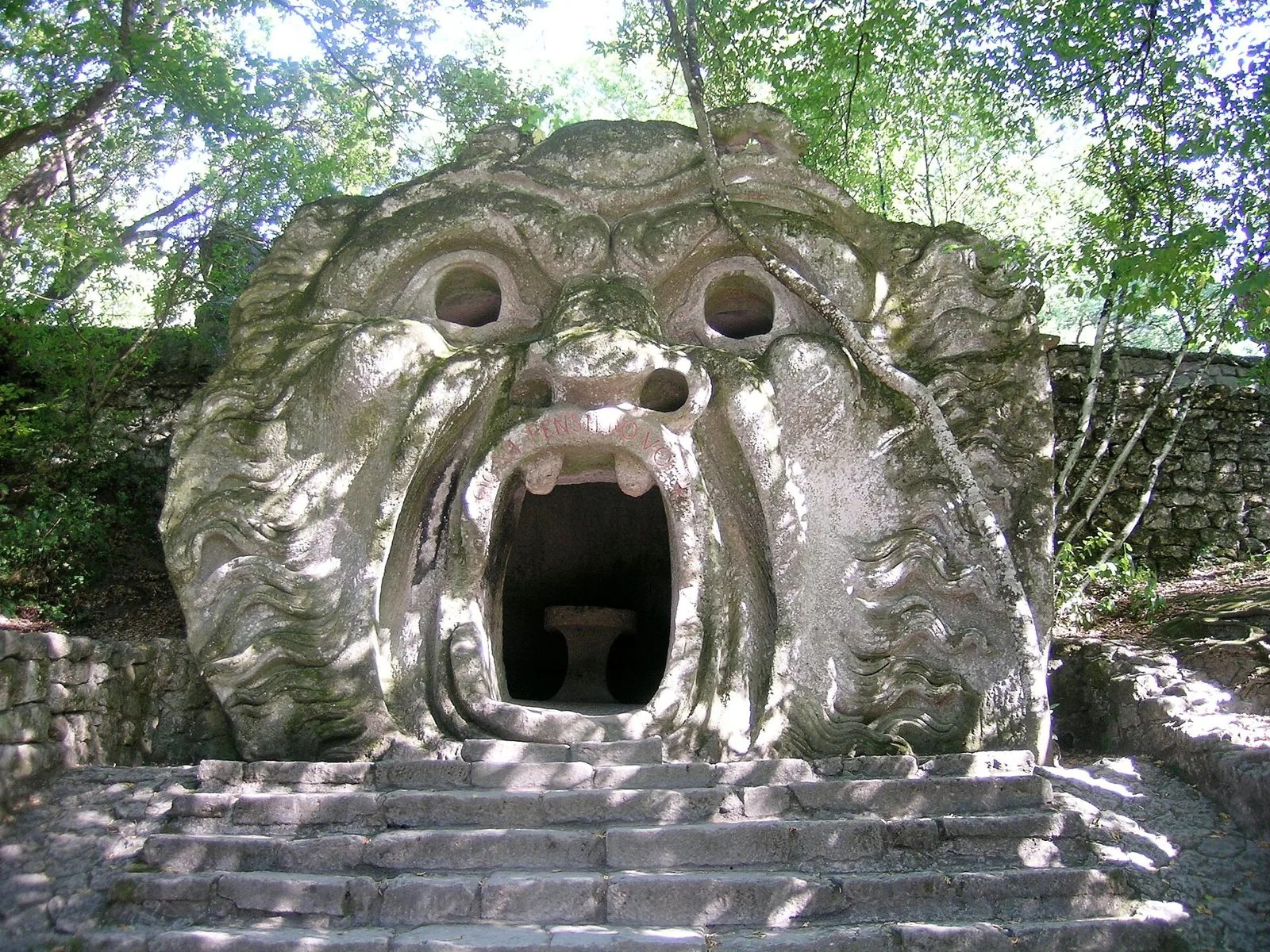 La scultura dell'Orco nel Sacro Bosco di Bomarzo, un volto gigante in pietra con la bocca spalancata e una scalinata d'accesso tra gli alberi
