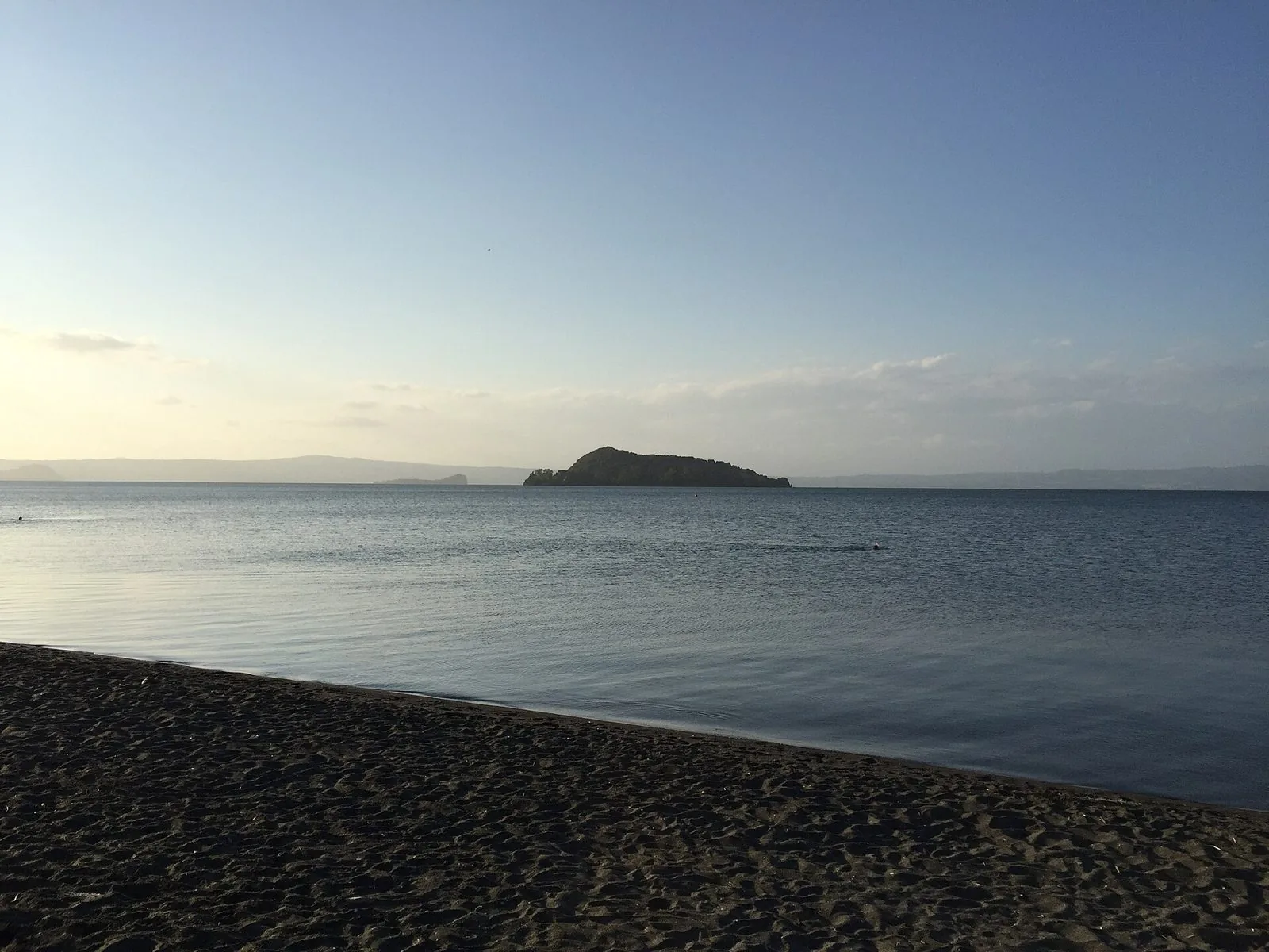 Spiaggia di sabbia scura sul Lago di Bolsena con l'acqua calma e un'isola verde all'orizzonte